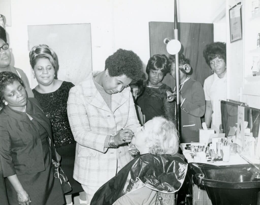 A group of ladies watch intently while one woman does another's makeup. 