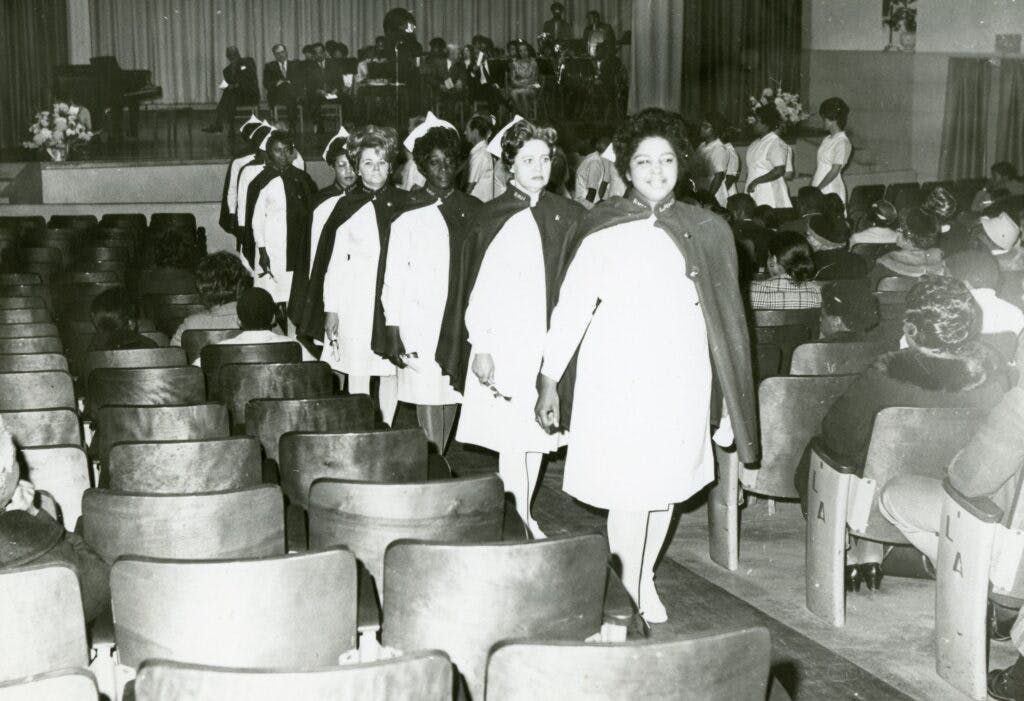 An auditorium with nurses dressed in their graduation capes and nurses uniforms. There is a band on stage and some audience members filling the seats