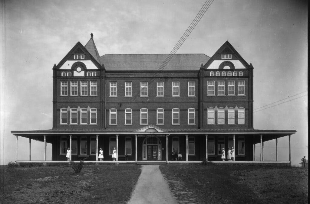 Black and white photo of Burrell Memorial Hospital. It has a highly semetrical design with a wrap-around awning. Nurses are pictured in all white dresses in the front of the hospital.