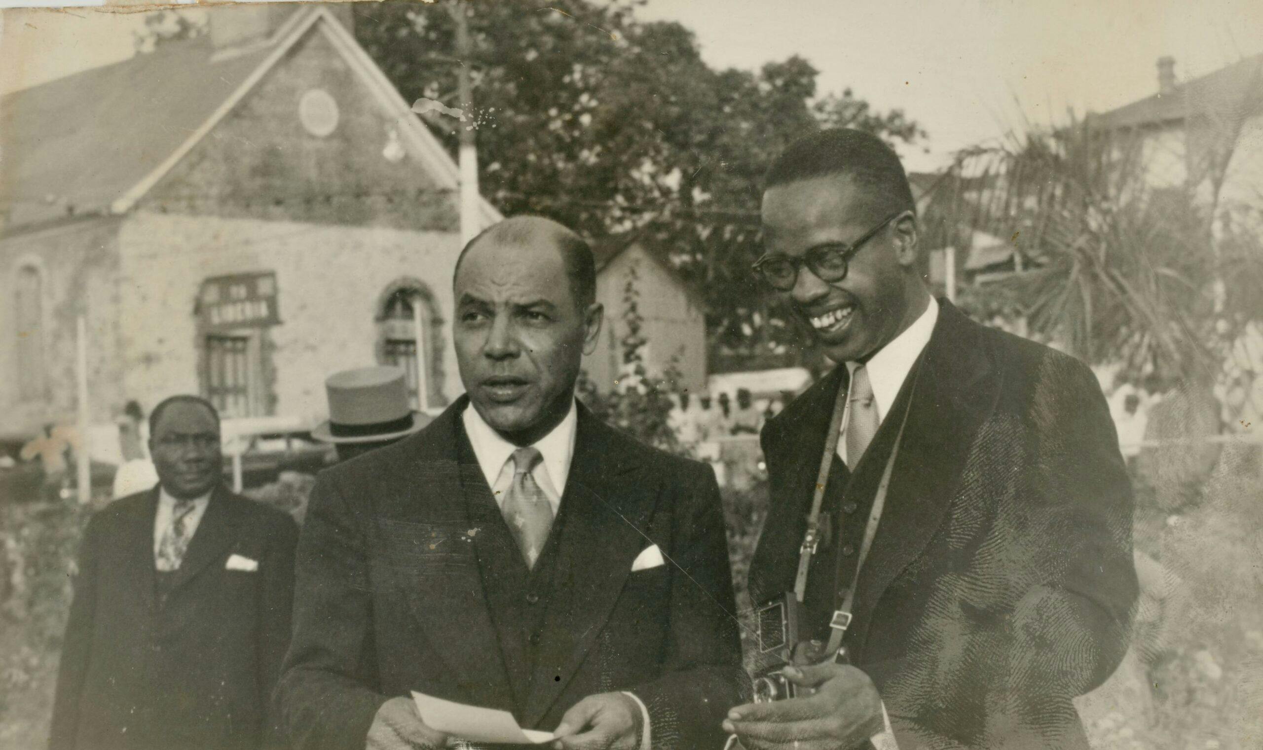 Old Photo of Ambassador Dudley with a photographer next to him. He is shown in a suit looking into the distance