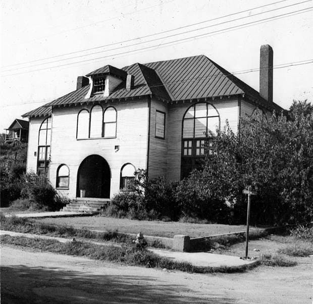 Black and white photo of the Gregory School, with its chimney and large front entrance and windows. It sits just off of a street with a short sidewalk