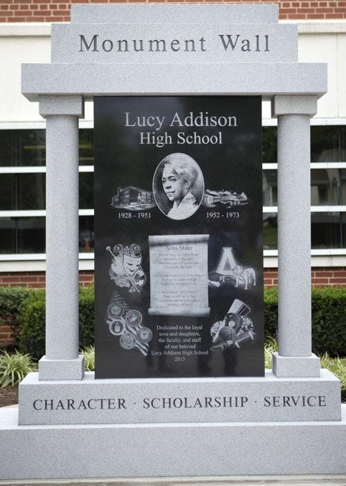 A monument wall carved in stone with Lucy Addison's portrait in the middle, flanked by both versions of the Lucy Addison High School building. Below that, there are several images: one for art, the Addison logo and mascot, an american flag with pins, sports, and the school's Alma Mater