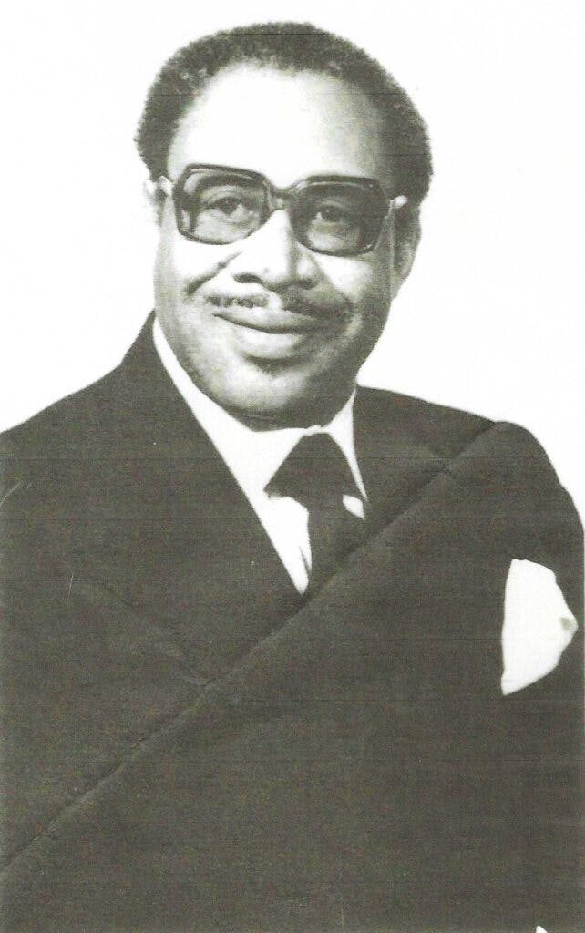 Reverend R.R. Wilkinson in a suit and tie and square framed eyeglasses.
