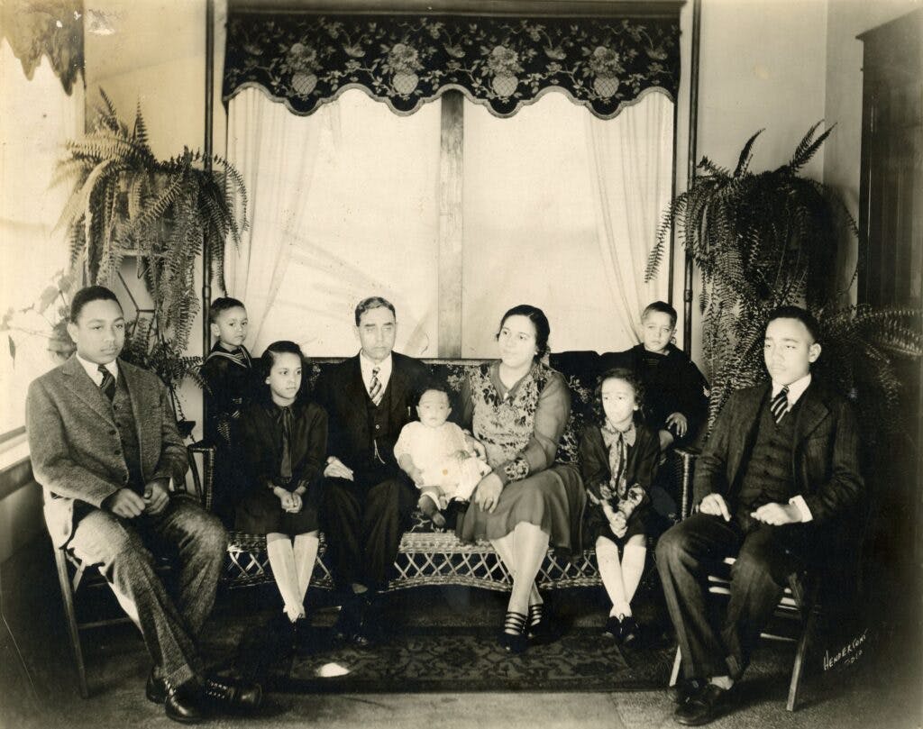 9 members of the Claytor family sit in their home with plants and a big window behind them